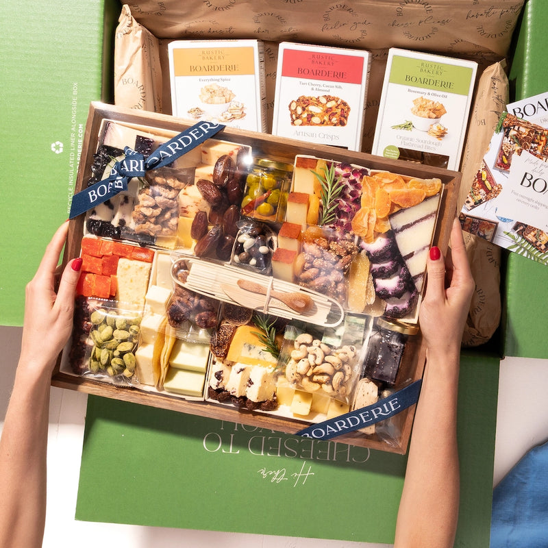 Gourmet cheese and charcuterie gift set showing wooden board with assorted cheeses, cured meats, nuts, olives, and Boarderie branded cracker boxes.