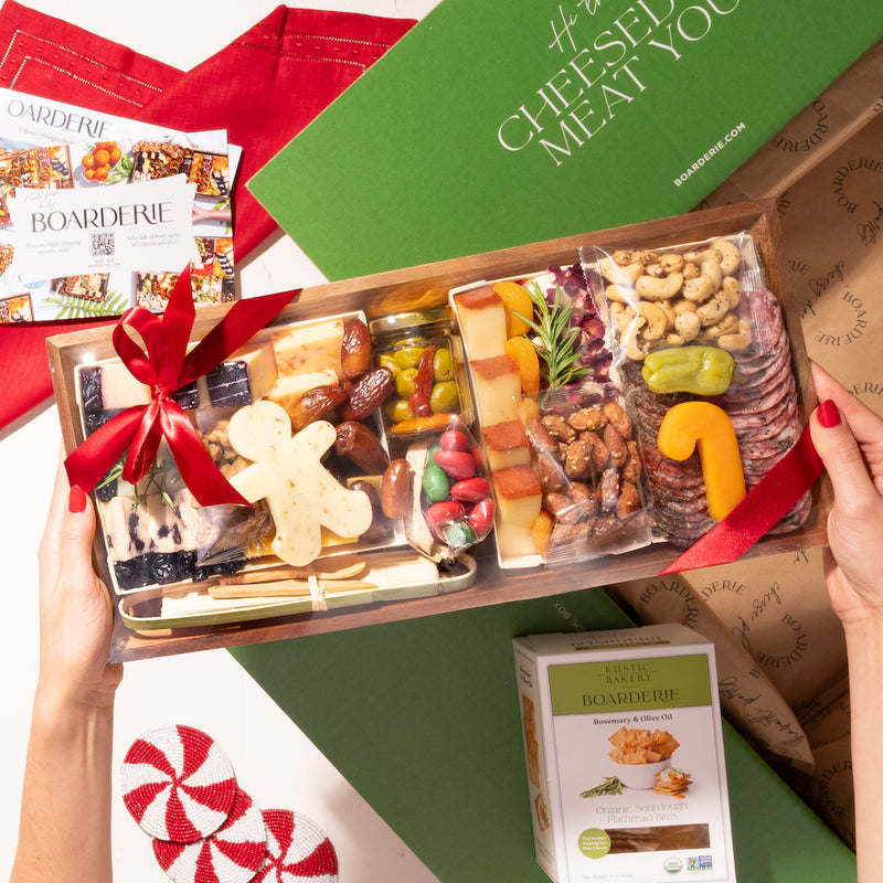 Holiday-themed small cheese and charcuterie board with festive cheese shapes, nuts, dried fruits, and crackers, tied with a red ribbon and boxed with artisan crisps