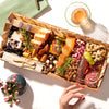 medium size charcuterie board with assorted cheeses, meats, nuts, and fruits, alongside a drink and decorative flowers.