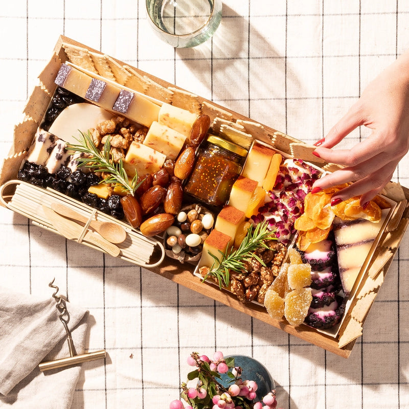 charcuterie board gift with an assortment of cheeses, dried fruits, nuts, crackers, dates, fig jam, & decorative rosemary sprigs