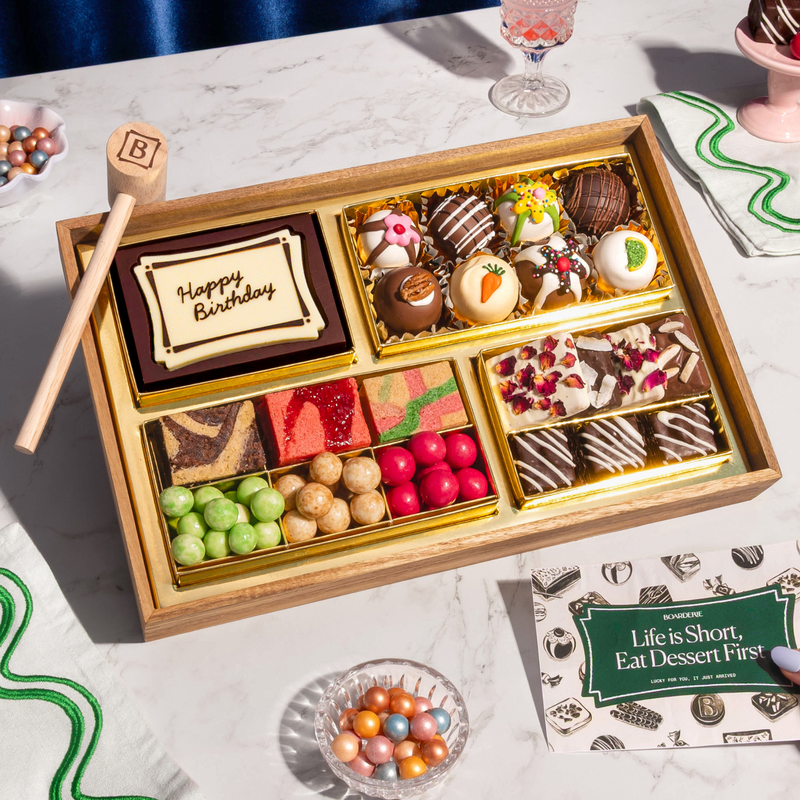 Small dessert gift box with "Happy Birthday" chocolate card, decorated bonbons, cake slices, chocolate-covered berries, and candied fruits on marble surface