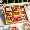 Dessert gift box featuring two rows of colorful macarons, decorated chocolate truffles, a "Thank you" chocolate plaque, brownies, rose petal bark, and candy balls in a gold-lined wooden tray