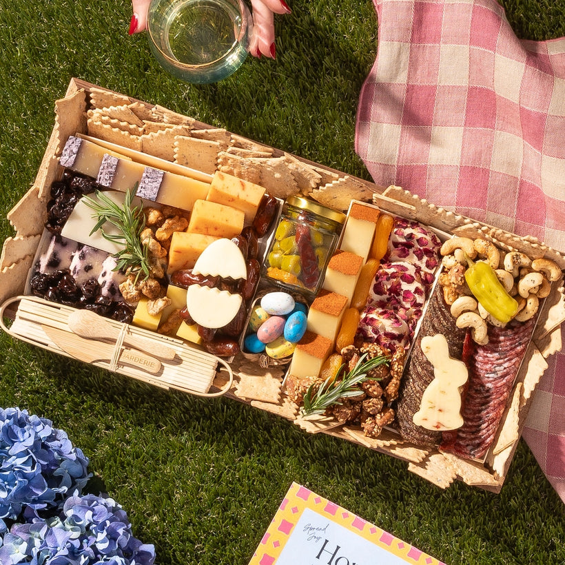 Easter egg charcuterie board with cheeses, meats, crackers, nuts, and festive egg and bunny-shaped treats on a picnic setting.