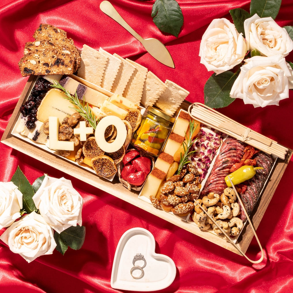 Romantic charcuterie board with "L+O" in cheese, surrounded by roses, rings, and a heart dish on red fabric.