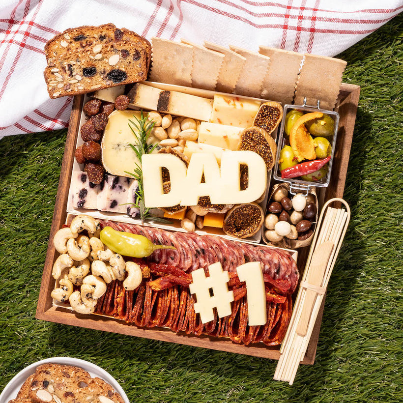 Father's Day charcuterie board with cheeses spelling "DAD" and "#1," meats, nuts, crackers, and olives on grass with a picnic cloth.