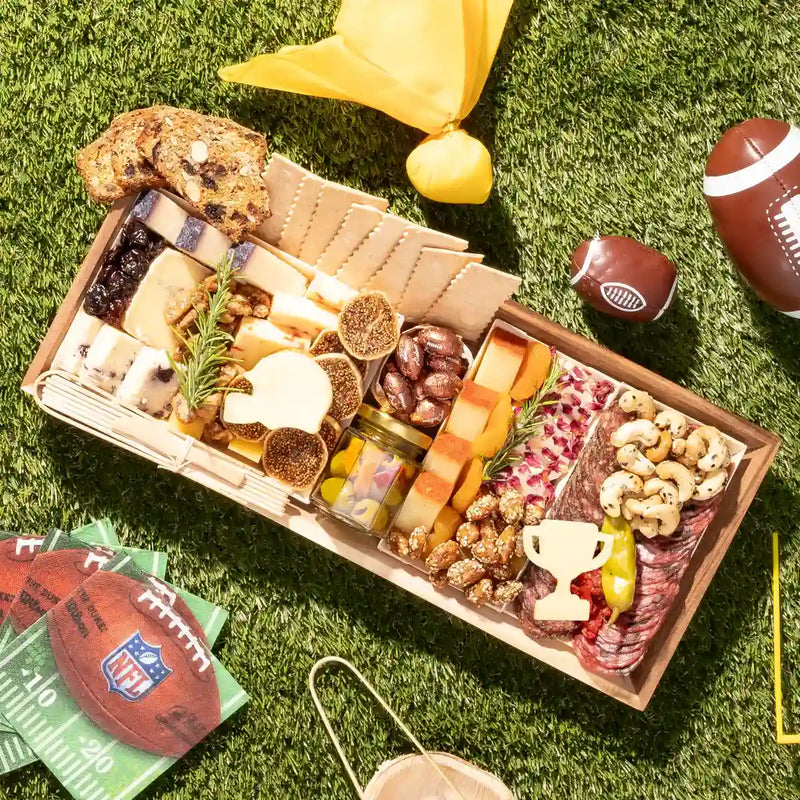 Football charcuterie board featuring cheeses, meats, crackers, nuts, and football-themed cheese cutouts on a picnic setting.