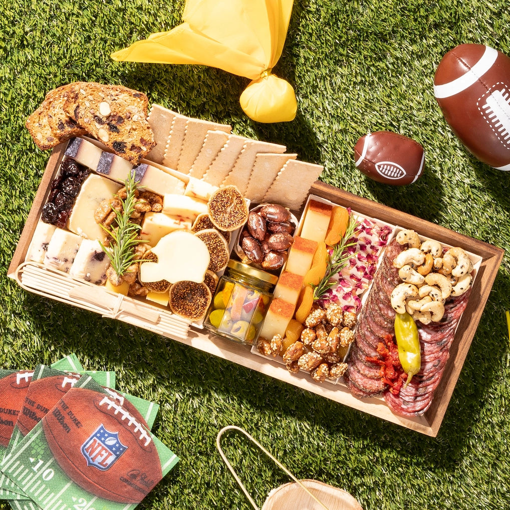 Football-themed charcuterie board with cheeses, meats, nuts, and a helmet-shaped cheese, set on grass with mini footballs and NFL napkins.