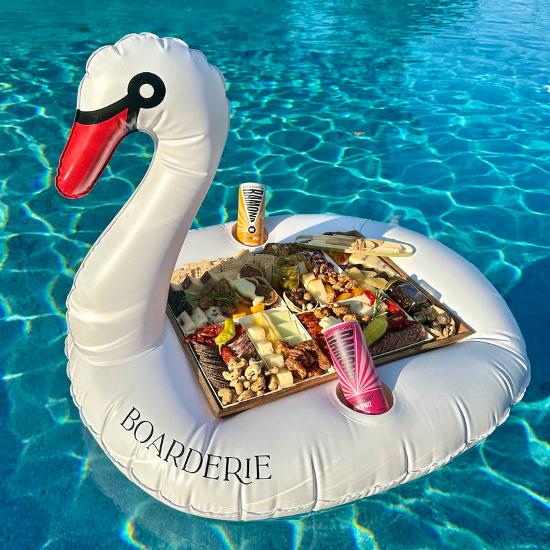 Boarderie charcuterie board floating on an inflatable swan in a pool, paired with canned beverages