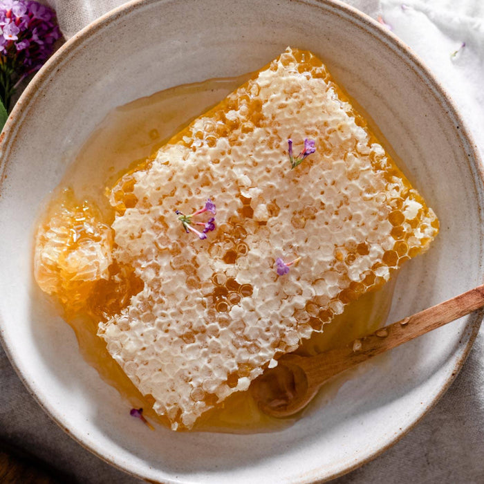 Fresh honeycomb with honey in cream bowl, garnished with edible purple flowers and wooden honey dipper on white background.
