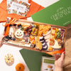 Halloween-themed medium size charcuterie board in festive packaging, featuring cheeses, nuts, and holiday decorations, held above a green box with crackers