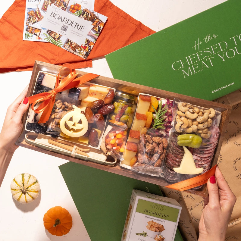 Halloween-themed medium size charcuterie board in festive packaging, featuring cheeses, nuts, and holiday decorations, held above a green box with crackers.