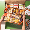 Halloween-themed charcuterie board in festive packaging, featuring cheeses, nuts, and holiday decorations, held above a green box with crackers.