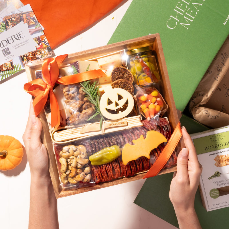 small halloween-themed charcuterie board in festive packaging, featuring cheeses, nuts, and holiday decorations, held above a green box with crackers