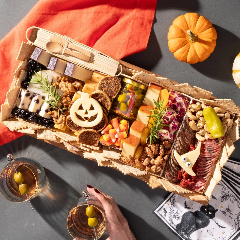 medium halloween charcuterie board featuring festive cheese shapes, candy corn, meats, and nuts, with pumpkins and cocktails nearby.