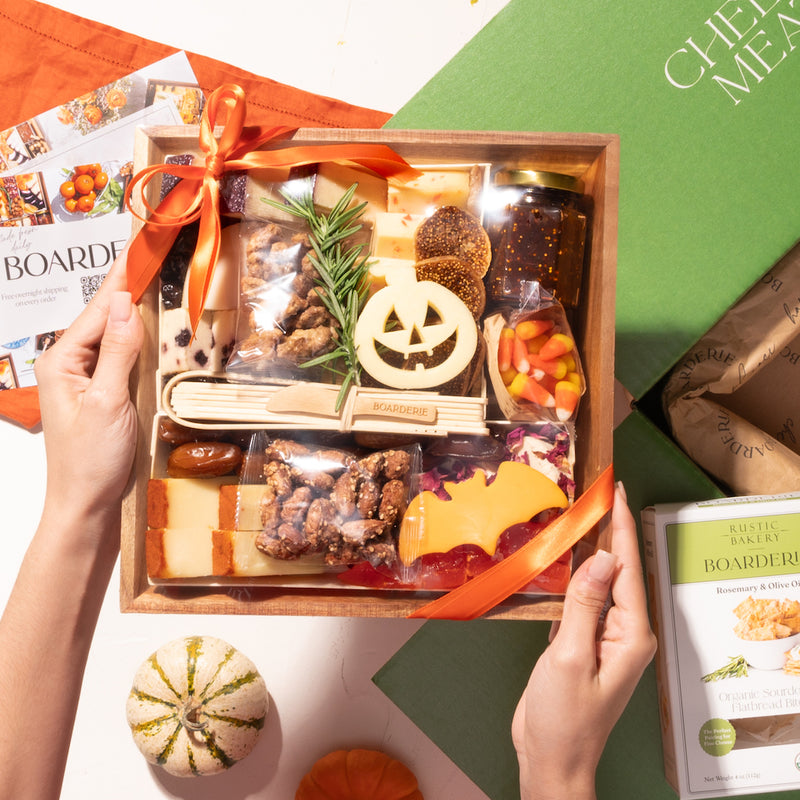 small halloween-themed cheese board in festive packaging, featuring cheeses, nuts, and holiday decorations, held above a green box with crackers