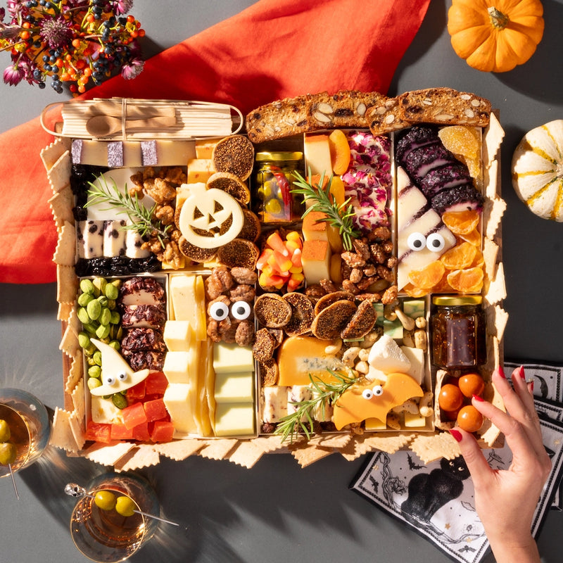 Halloween-themed charcuterie board featuring festive cheeses shaped like a pumpkin and bat, surrounded by assorted snacks and decorations.