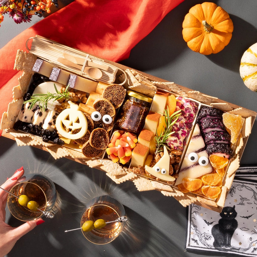 medium size halloween charcuterie board featuring festive cheese shapes, candy corn, meats, and nuts, with pumpkins and cocktails nearby