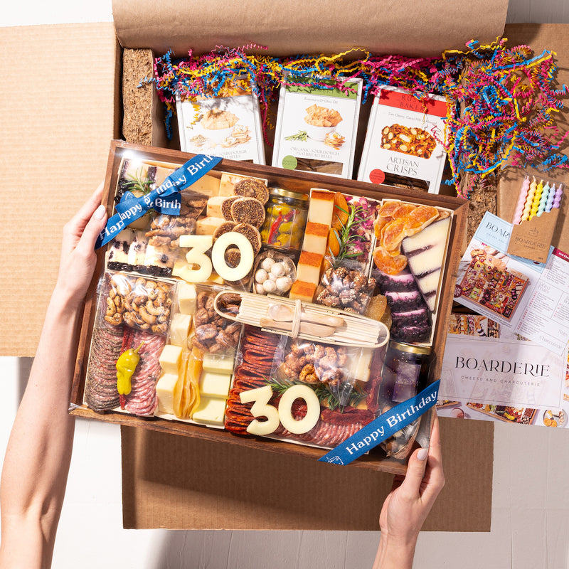 Hands holding a birthday charcuterie box with "30" in cheese, wrapped in blue "Happy Birthday" ribbon and colorful confetti.