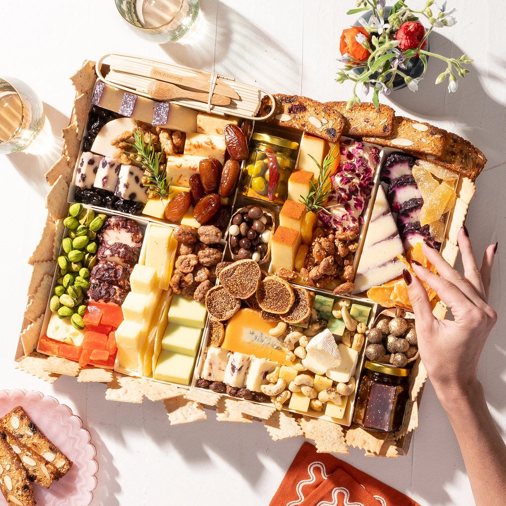 Large cheese board with assorted cheeses, nuts, dried fruits, and crackers, styled with flowers and wine on a white table.