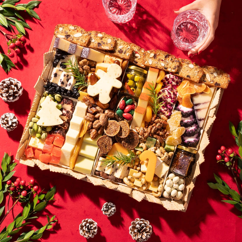 Large Christmas-themed cheese board with assorted cheeses, nuts, dried fruits, & festive decorations on a red tablecloth