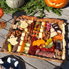 A Halloween-themed charcuterie board arranged on a wooden table, featuring sliced meats, cheeses, nuts, dried fruit, candy, and spooky decorations like a ghost cheese, bat cheese and jack-o'-lantern cheese