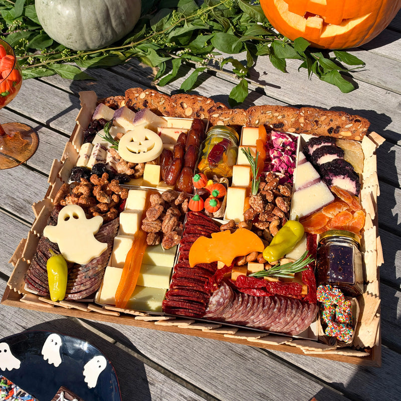 A Halloween-themed charcuterie board arranged on a wooden table, featuring sliced meats, cheeses, nuts, dried fruit, candy, and spooky decorations like a ghost cheese, bat cheese and jack-o'-lantern cheese