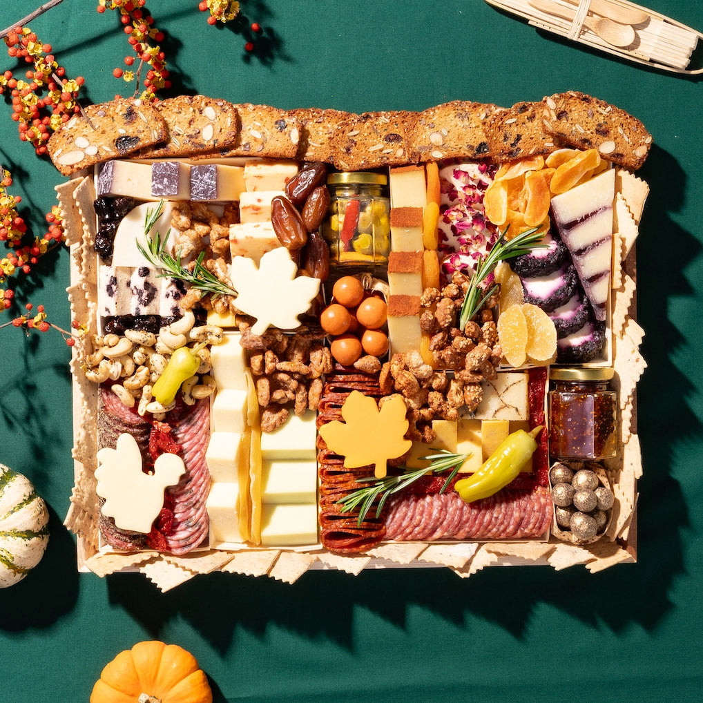 Thanksgiving charcuterie board for 9+ people with assorted cheeses, meats, crackers, dried fruits, and fall-themed decorations on a green tablecloth.