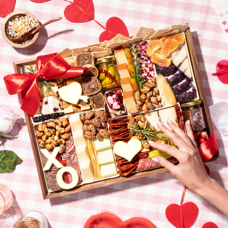 large Valentine's Day charcuterie board gift adorned with heart and "XO" shaped cheeses, assorted meats, nuts, dried fruits, crackers, & figs, wrapped with a festive red ribbon