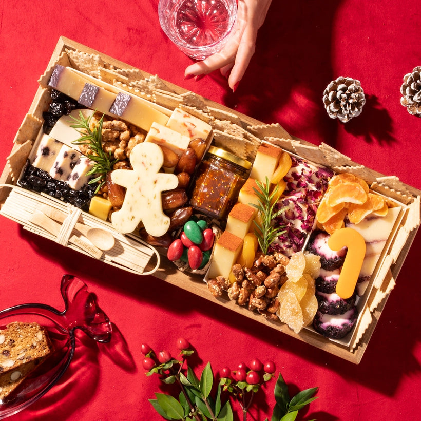 Medium holiday cheese board with festive cheese shapes, nuts, dried fruits, fig jam, and crackers on a red tablecloth.