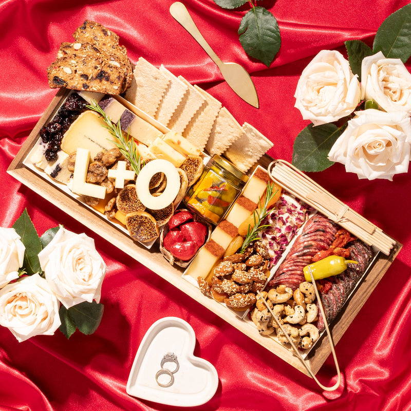 Romantic charcuterie board with "L+O" in cheese, surrounded by white roses on a red satin background, with wedding rings in a heart dish.