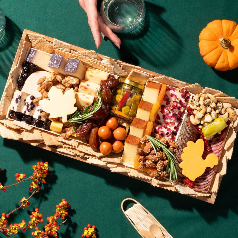 medium thanksgiving charcuterie board with assorted cheeses, meats, nuts, & fall-themed decorations on a green tablecloth