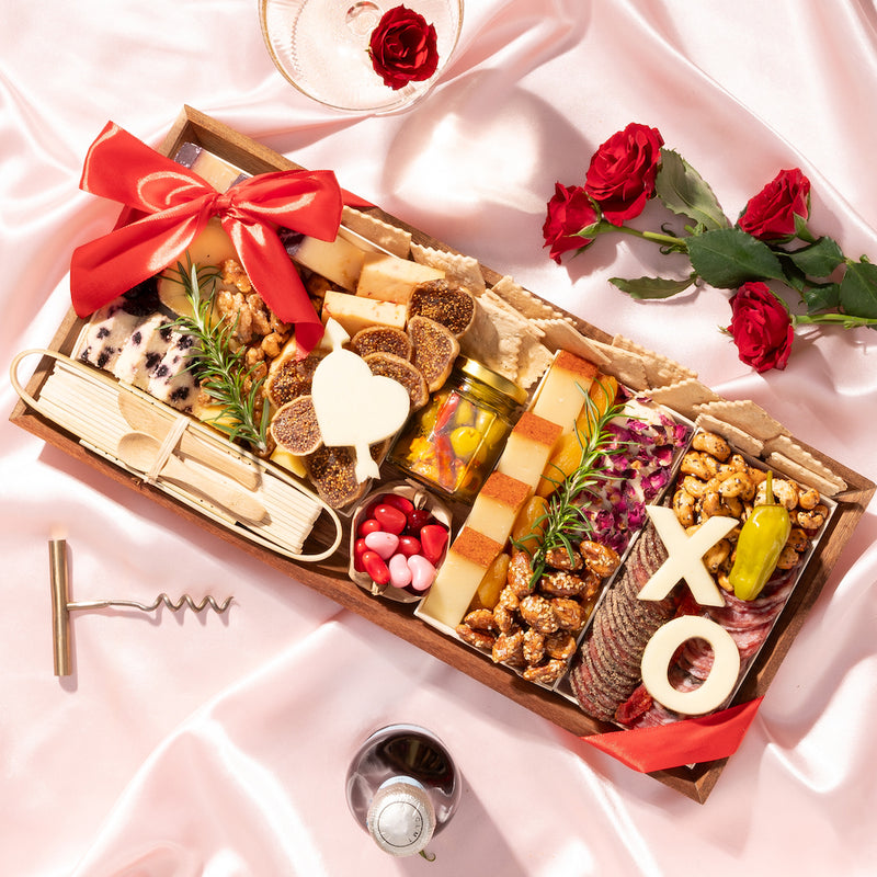 medium Valentine's Day charcuterie gift board featuring heart and "XO" shaped cheeses, nuts, crackers, cured meats, figs, & a red ribbon