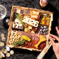 Wooden New Years charcuterie board displaying assorted cheeses, cured salami slices, roasted cashews, marinated olives, berry jam, dark chocolate pieces, and decorative 2026 numeral with fresh rosemary sprigs.