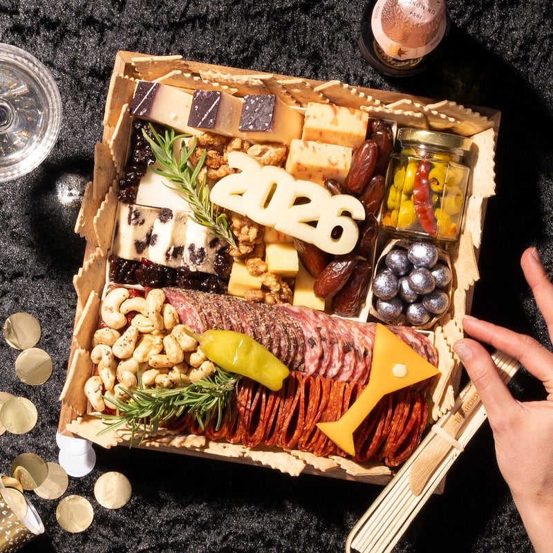 Wooden New Years charcuterie board displaying assorted cheeses, cured salami slices, roasted cashews, marinated olives, berry jam, dark chocolate pieces, and decorative 2026 numeral with fresh rosemary sprigs.