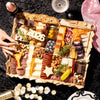 Large New Years party charcuterie board with wooden frame featuring assorted cheeses, cured meats, nuts, olives, star-shaped cutouts, and 2026 number decoration surrounded by champagne and candles.