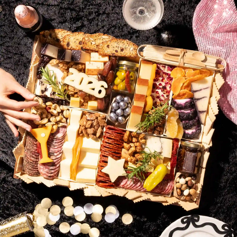 Large New Years party charcuterie board with wooden frame featuring assorted cheeses, cured meats, nuts, olives, star-shaped cutouts, and 2026 number decoration surrounded by champagne and candles.