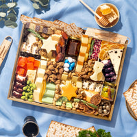 Large Passover cheese board overflowing with multiple Star of David cheese cutouts, assorted artisan cheeses, candied nuts, dried apricots, sliced figs, fig jam, blue and white candies, and matzo on a blue linen surface