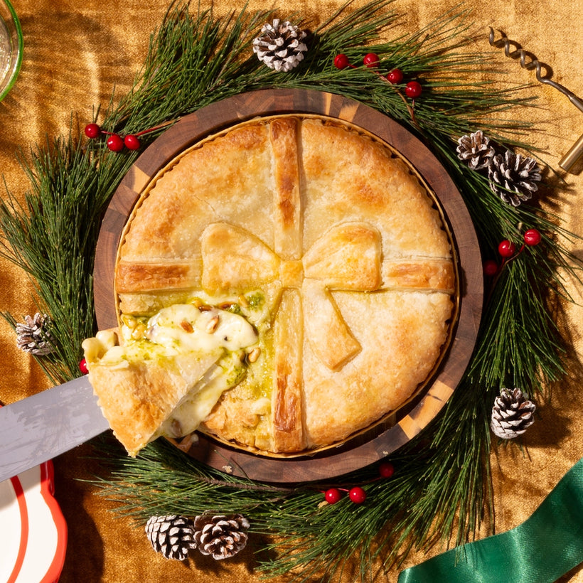 Holiday baked brie shaped like a gift with melted cheese and pine nuts, surrounded by festive pine and berries.