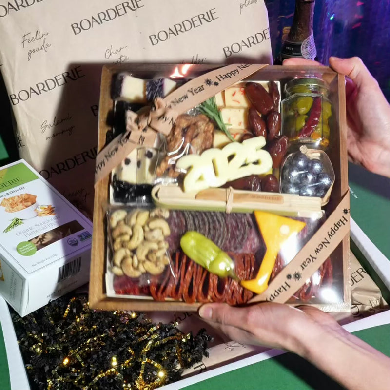 video of New years eve charcuterie board in front of box then shown unwrapped with artisan cheeses, meats, nuts, fried fruits, chocolates and olives on a wood board