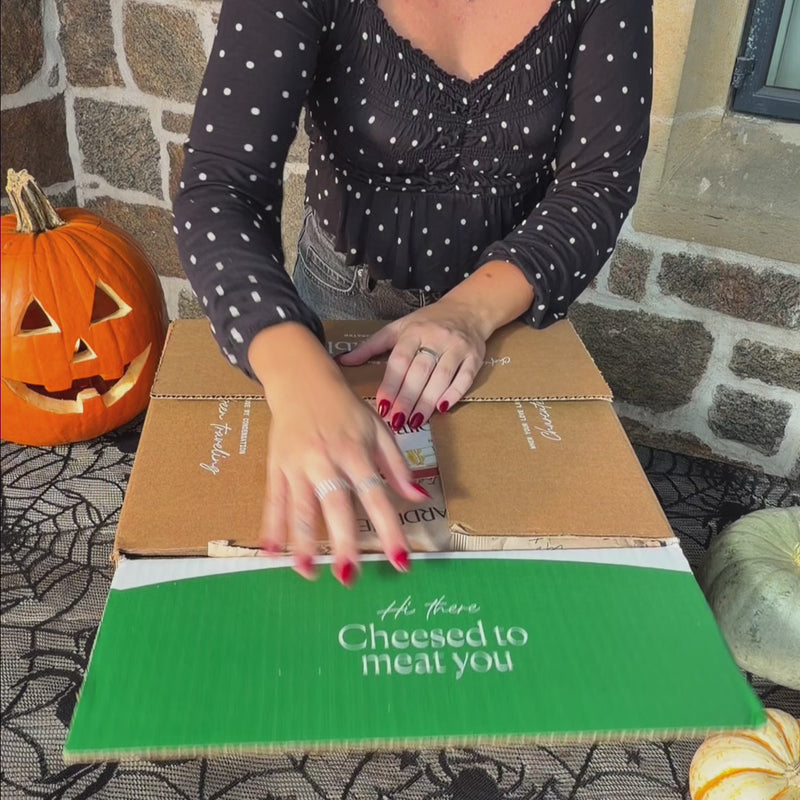 Unboxing of a Halloween-themed charcuterie board with jack-o'-lantern cheese cutout, googly-eyed candy treats, cured meats, nuts, olives and more.