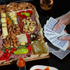 Large charcuterie board with assorted cheeses, meats, nuts, crackers, and peppers, set beside a card game and wine glass.