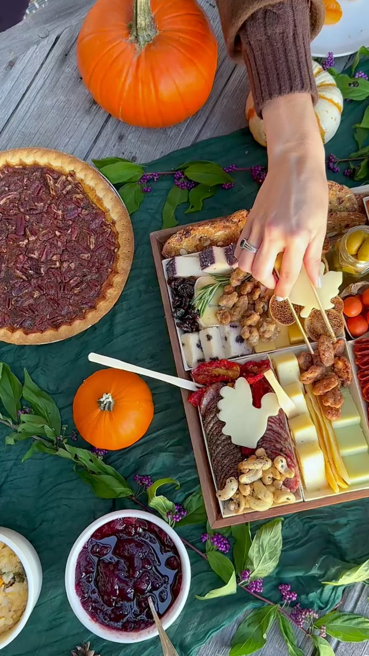 Thanksgiving appetizer spread with the cheese and charcuterie board centerpiece and hands picking up nuts, cheese, and meats.