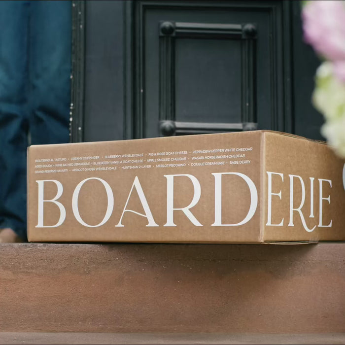 Boarderie box at front step, unboxing of cheese and charcuterie board classic, a couple smiling and clinking glasses together, nuts pouring onto board, girl with blonde hair smiling at board, hands picking at nuts and cheeses on board, Christmas themed board