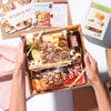 Hands holding a neatly packed charcuterie box with assorted cheeses, meats, nuts, & figs, next to Boarderie pamphlets.