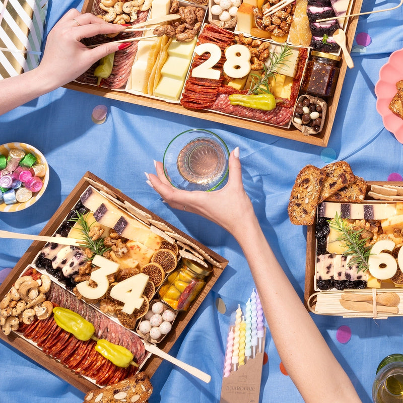 Birthday charcuterie boards with numbers "28," "34," and "60" in cheese, surrounded by assorted snacks on a festive blue tablecloth.