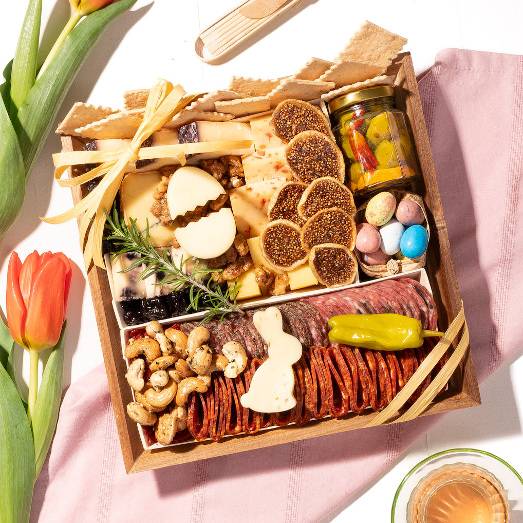 Easter-themed charcuterie board with cheese, meats, nuts, pastel eggs, and decorative egg and bunny cheese shapes, accented with tulips.