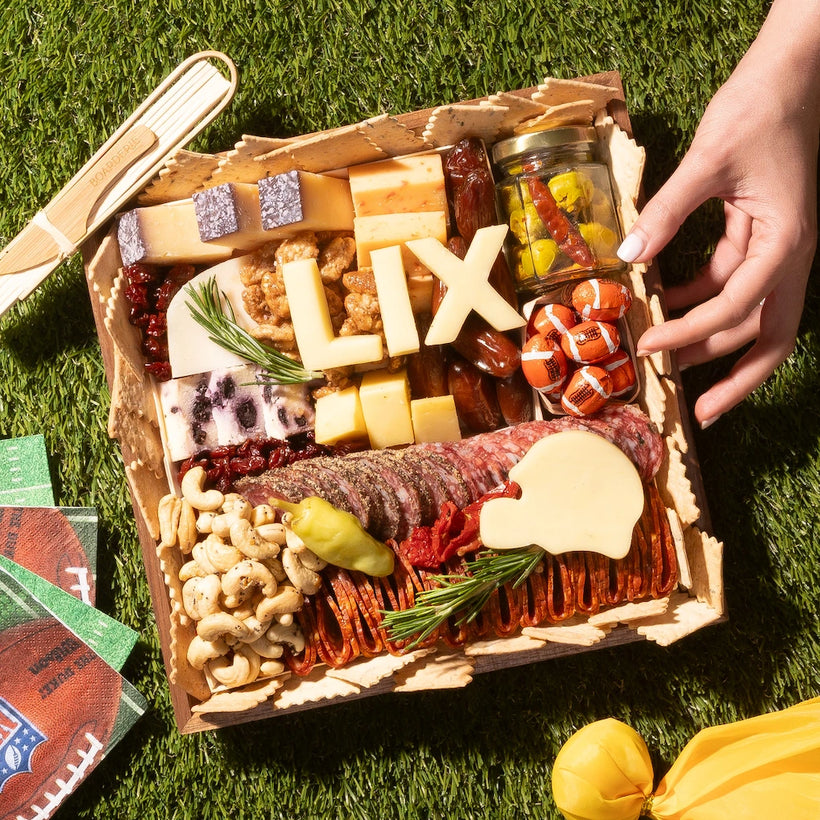 Super Bowl-themed charcuterie board with cheeses, meats, crackers, nuts, and football-inspired cheese cutouts on a grass picnic setting.