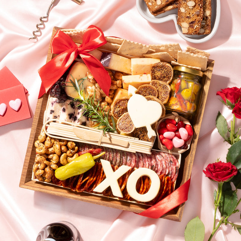 small Valentine's Day charcuterie gift board decorated with heart and "XO" shaped cheeses, assorted meats, nuts, figs, & crackers, accented with a red ribbon