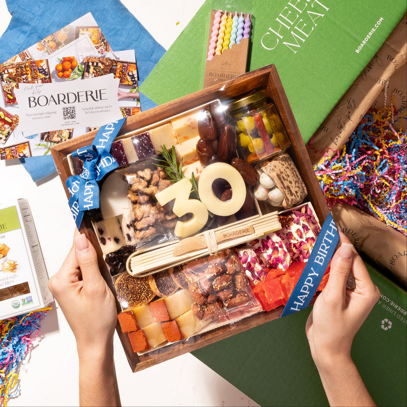Birthday Cheese Board being unboxed, fully arranged gourmet birthday gift with 19 artisan cheeses, dried fruits, nuts, chocolates, jam and crackers on a reusable acacia wood board. Includes bamboo utensils, birthday candles, confetti and customizable age numbers in cheese. Option to add personal gift note. Ships free overnight nationwide in insulated packaging with next-day delivery available. Perfect for birthday gifts, corporate gifting and party hosting.