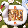 Charcuterie board arranged on a swan-shaped float with various snacks and drinks, set on grass for an outdoor picnic